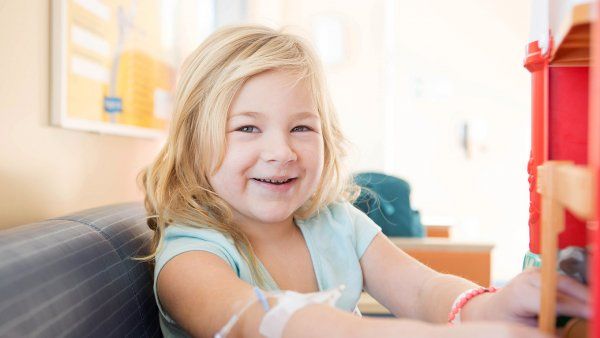 Child smiling in a hospital chair, with a medical line in her arm.