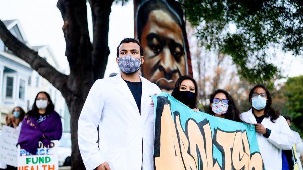 UCSF Nurses gather in Oakland for a Black Health Matters protest; a male Black nurse in a white coat and face mask stands with female nurses, holding a sign that reads “Abolish” in graffiti style; a large mural of George Floyd’s portrait is in the background.