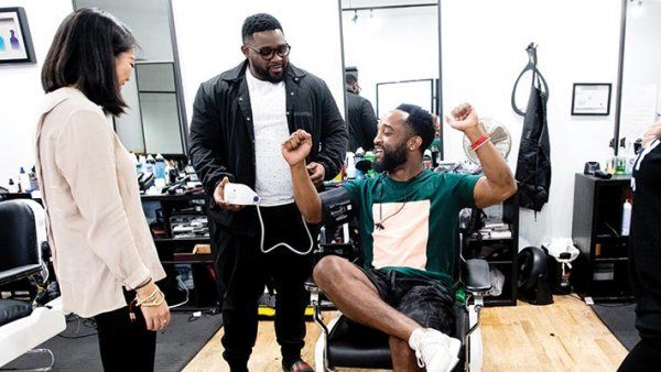 Photo of barbers Joseph Peters, seated in a barber’s chair, and Terell Kennedy standing behind the chair, with two women standing in a barber shop.