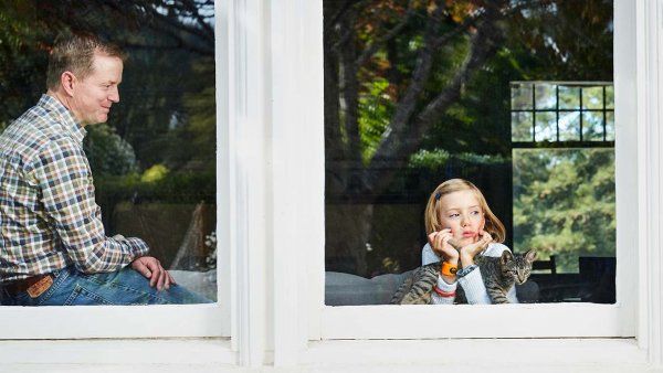 Tim Wood looks down at his daughter, Piper; Piper is holding a cat and looking out the window.