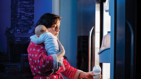 Photo of a tired mother with a newborn baby, reaching for a bottle of milk from the refrigerator at night.