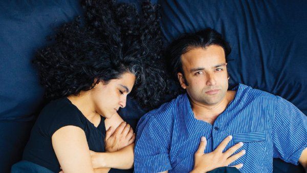 Photo of a man and woman in bed; the woman is fast asleep and the man eats popcorn, wide awake.