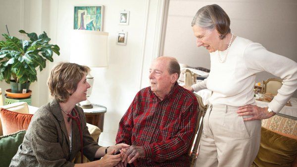 Dr. Rebecca Conant sits with an elderly male patient and an elderly woman stands behind him.