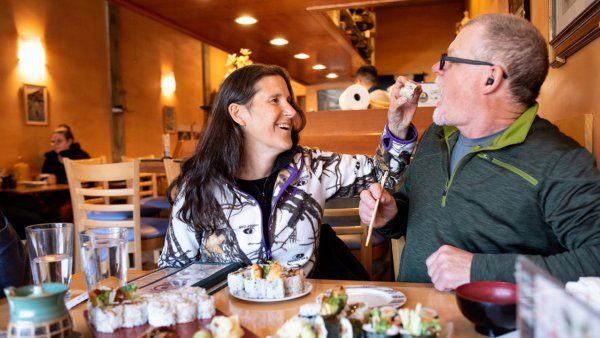 Cheryl feeds her partner sushi with a pair of chopsticks while both laugh