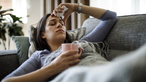 Sick woman lying on her couch, holding a mug and a tissue