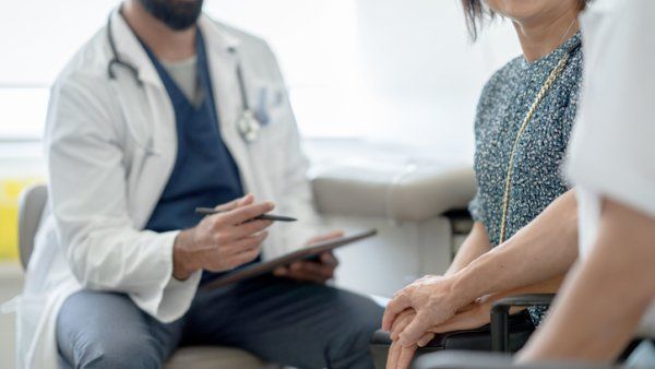 A senior couple hold hands to support one another as the doctor shares the news of the woman's test result with them. The doctor is wearing blue scrubs and a lab coat over top as he holds out a tablet in his hands and talks with the couple.