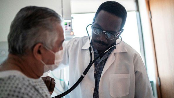 An elderly man wearing a mask and hospital gown sits on hospital bed as a doctor listens to his chest with a stethoscope