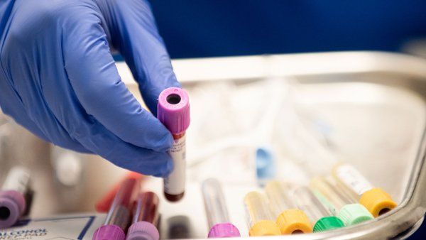 A hand in a blue latex glove holds a vial filled with blood