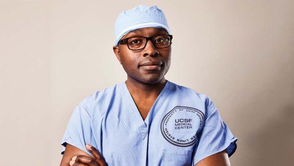 Portrait of neurosurgeon Shawn Hervey-Jumper, standing with his arms crossed in blue surgical scrubs.