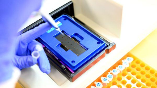 A researcher injects liquid from vials into a device. Next to the device is an orange tray with vials.