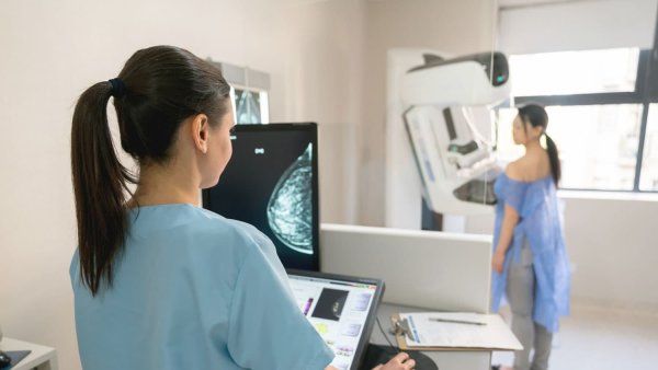 A technician takes a mammogram. The woman getting the mammogram stands at the far end of the room. The scan can be seen on the computer screen.