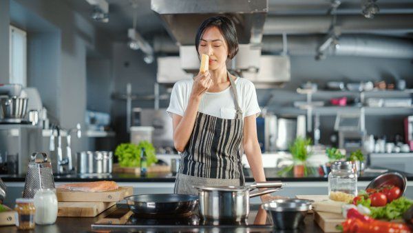 A female chef holds a slice of cheese as she sniffs it. She stands in a large kitchen