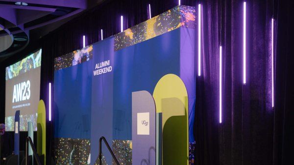 The stage of Alumni Weekend showing long purple hanging lights, a purple backdrop, and a podium