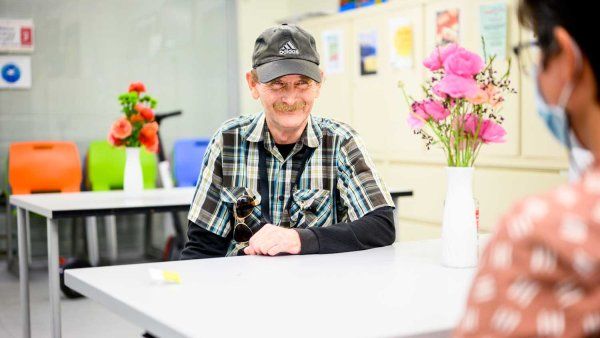 Bill smiles as he sits at a table and speaks with a social worker. He wears a cap and a plaid shirt.