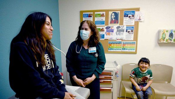 A female Hispanic teenager sits on an examination table as she speaks with a female doctor. Her younger sister sits in the background.