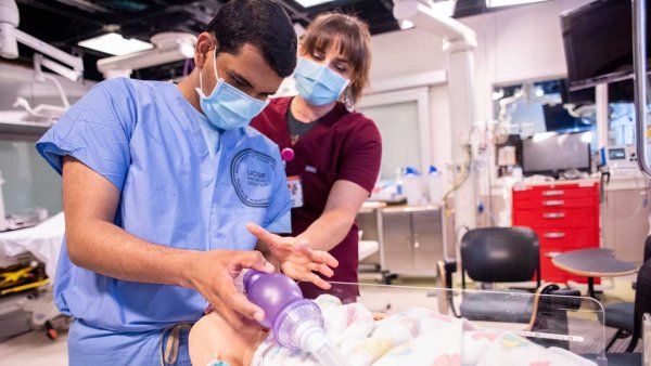 A medical student wearing blue scrubs practices delivering oxygen to a dummy while a doctor in red scrubs provides instruction.