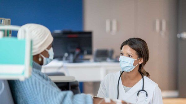 A woman wearing a beanie and surgical mask sits on a chair as she receives chemotherapy. A nurse kneels as she explains a procedure.