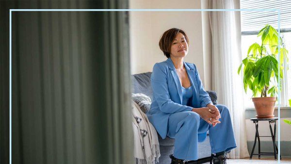 Caro reyes sits on a couch in her brighlty lit living room. A green plant is in the background.