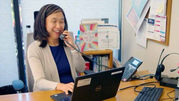 Miriam Rhew smiles as she consults with someone on a phone and looks at her laptop.