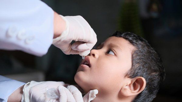 A pair of gloved hands deliver an oral polio vaccine with a dropper into a young boy's mouth.