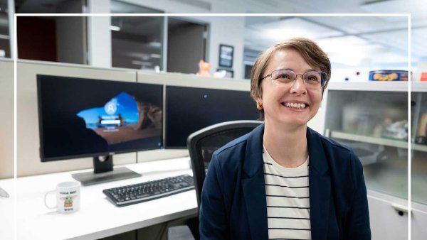 Mindy Hebert-DeRouen smiles as she sits in her chair at her work cubicle. Behind her are two computer screens on her desk.