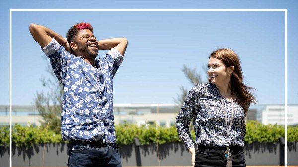 Chase Anderson stands outside under a blue sky and gleefully stretches his arms behind his head while he laughs. His colleague Caitlin stands behind him and smiles as she watches him.