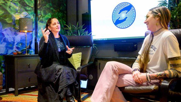 Jennifer Mitchell, left, speaks with a clinical reasearch coordinator, right, in a therapy room. Behind them is a screen, and the carpet is a colorful array of orange and yellow. 