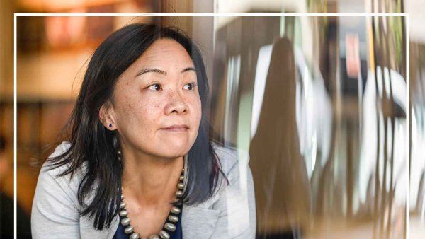 Angela Woon looks outside of a window. Reflected on the window are buildings on a San Francisco street.