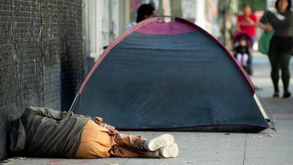 A homless person sleeps on a sidewalk next to a tent.