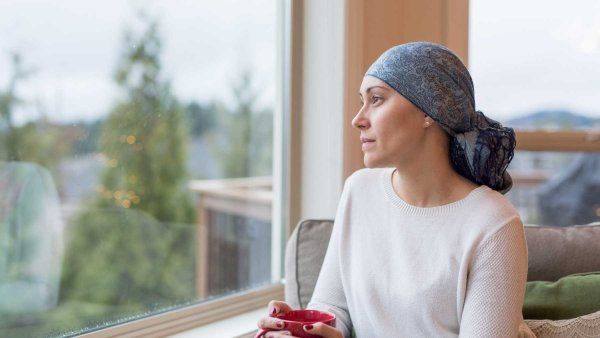 A young woman with cancer sits by her living room window and gazes out contemplatively. She is wearing a headscarf and drinking a cup of tea.