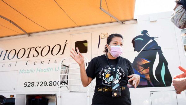 An indigenous woman wearing a surigcal mask stands outside a mobile clinic while talking to a patient in Ford Defiance.