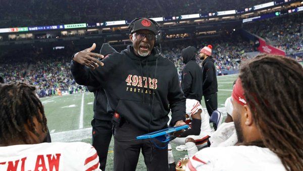 San Francisco 49ers coach Johnny Holland holds a tablet while coaching two 49ers players on the sidelines. Crowds fill stadium seats in the background.