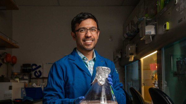 Aashish Manglik wears a blue lab coat and holds a large chemical flask with a pink liquid.