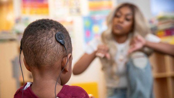 Amir, a toddler, wears two Cochlear implants on the back of his head. In the background, his mother Alisa communicates with him through sign language. He has a cochlear implant, as he became deaf after an infection. They are both of African American descent.