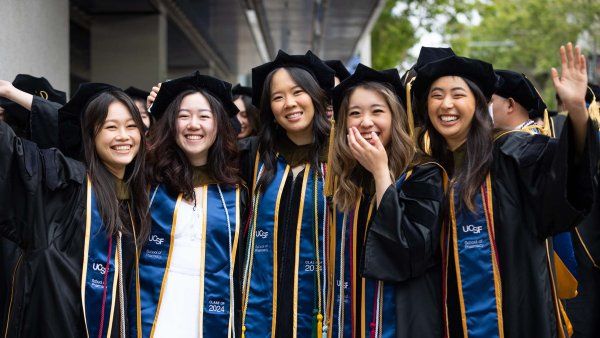 Five new School of Pharmacy graduates laugh and raise their arms outside of the ceremony hall.