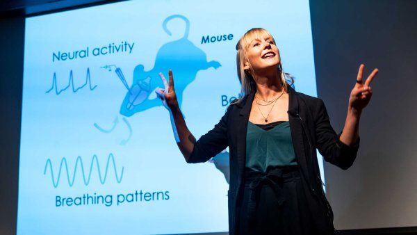 Alexandra Klein, the 2024 Postdoc Slam winner, stands onstage and delivers her talk. In the background is a projection screen with a darwing of a mouse, and measurments of brain activity.