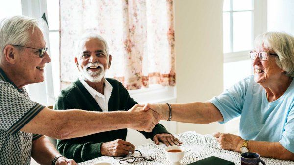 A group of three elerdly people smile together as they sit at a sun-lit kitchen.