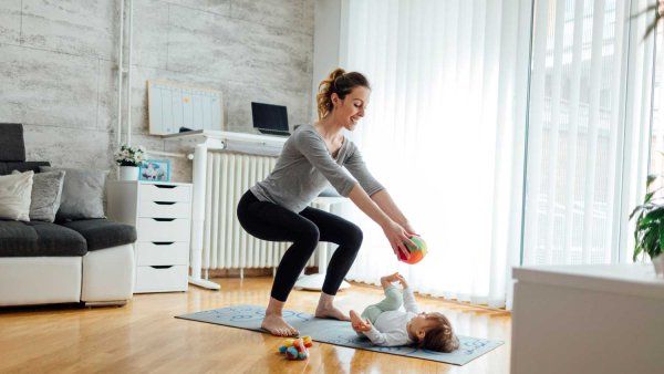A young mother does exercises on a yoga matt while her baby lays on its back in front of her.