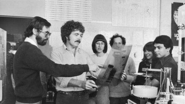 An archivial black and white photo of Herbert Boyer with staff, looking at radiogram of experiment.