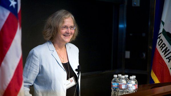 Elizabeth Blackburn smiles as she addresses a crowd behind a podium and in front of an American flag.