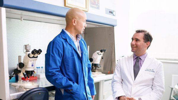 Johan Chan leans against a table while speaking with Ari Green (right) in a lab. Chan wears a blue lab coat while Green wears a white coat.